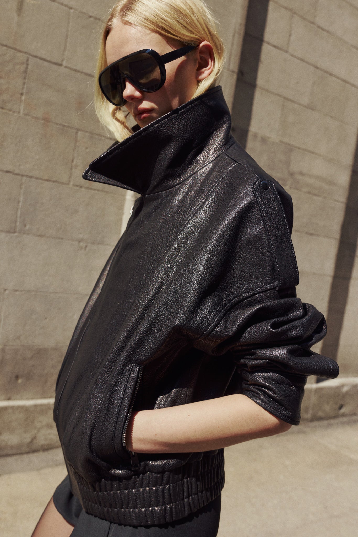 A blonde model wearing oversized black leather jacket with a high collar, photographed outdoors against a gray stone wall.
