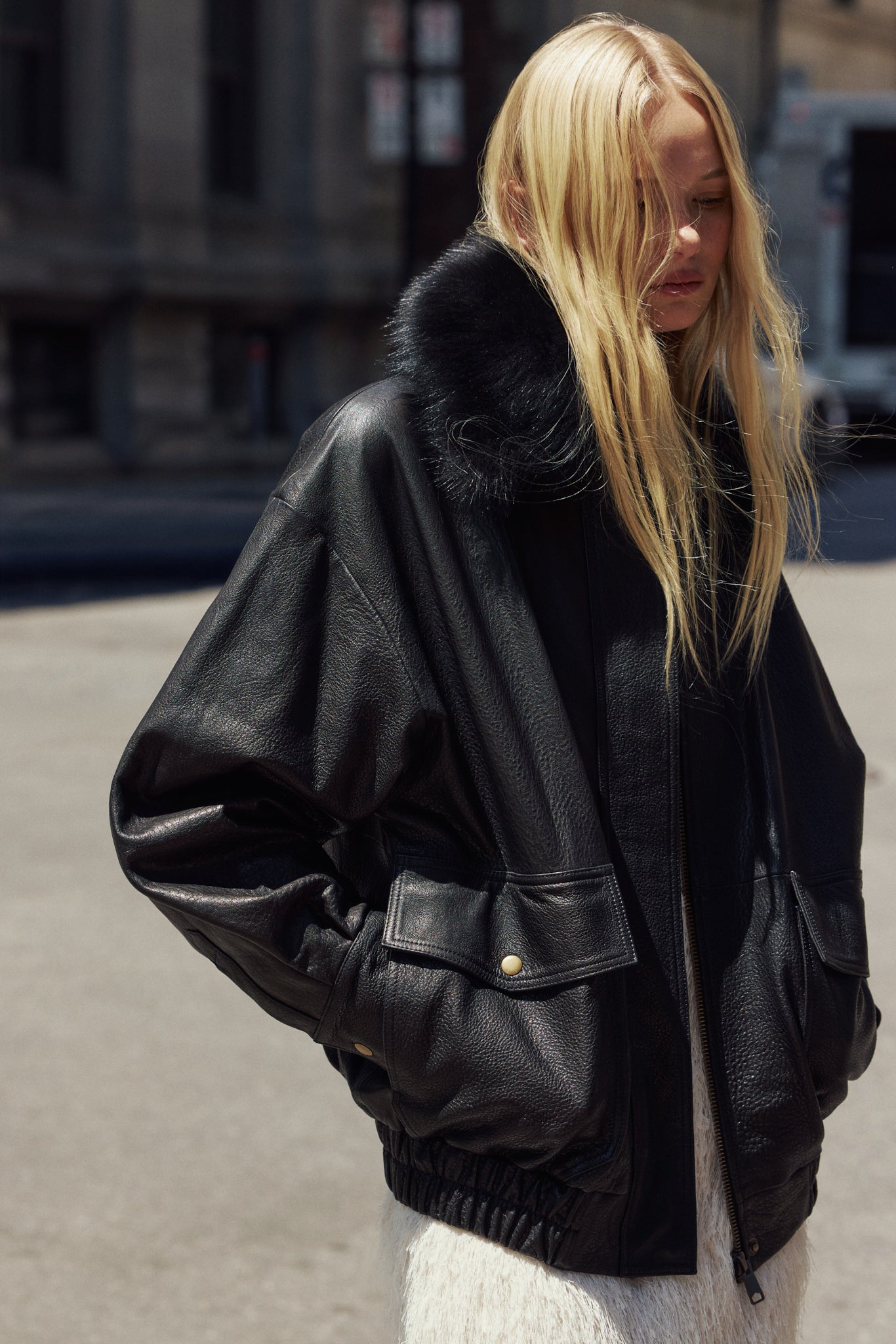 A blonde woman wearing TATE a black leather bomber jacket with a black shearling collar, standing outdoors on a city street.