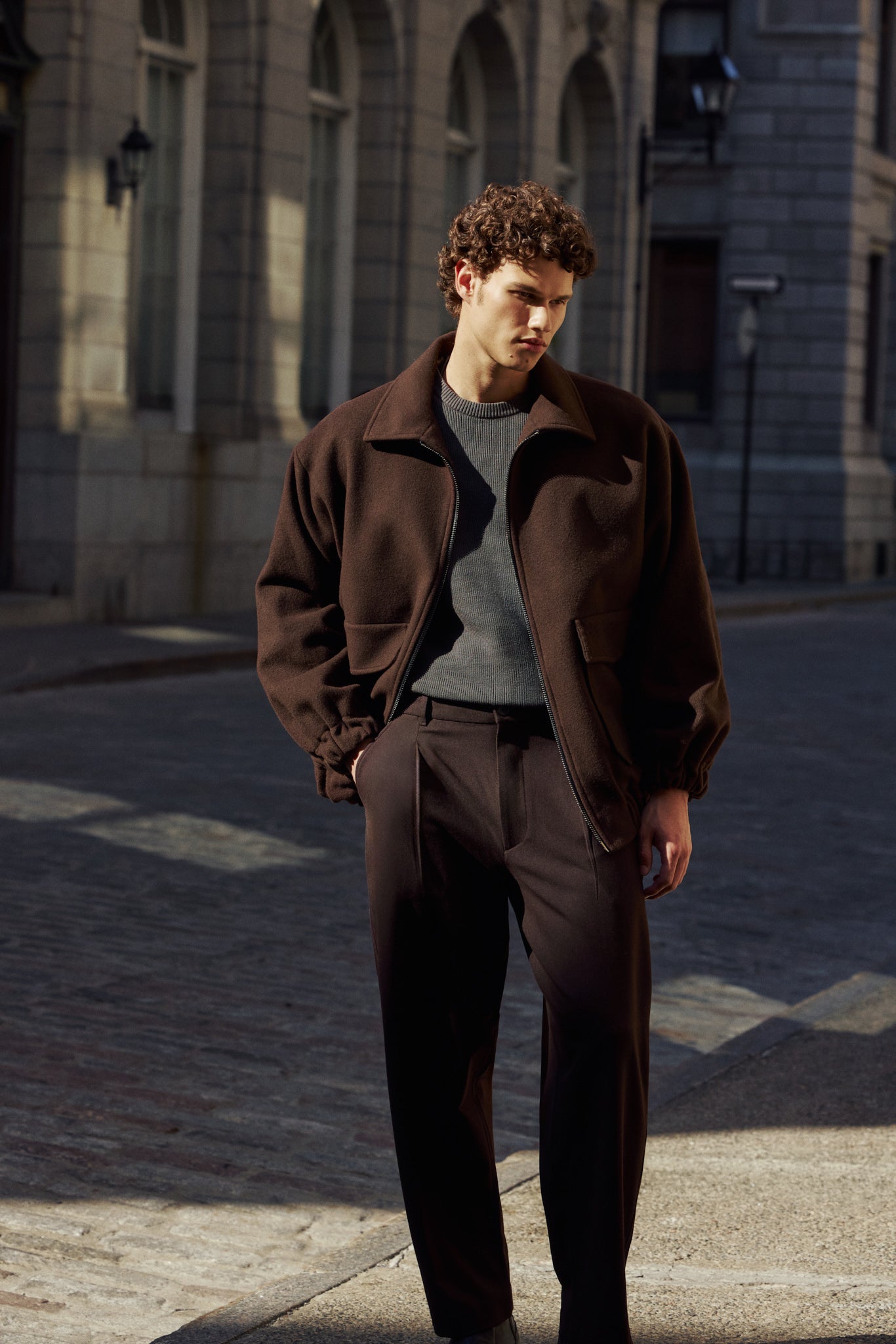 A man wearing a dark brown zip-up bomber jacket and matching dark brown trousers stands in profile against a light beige stone wall, with strong sunlight casting a shadow on the wall