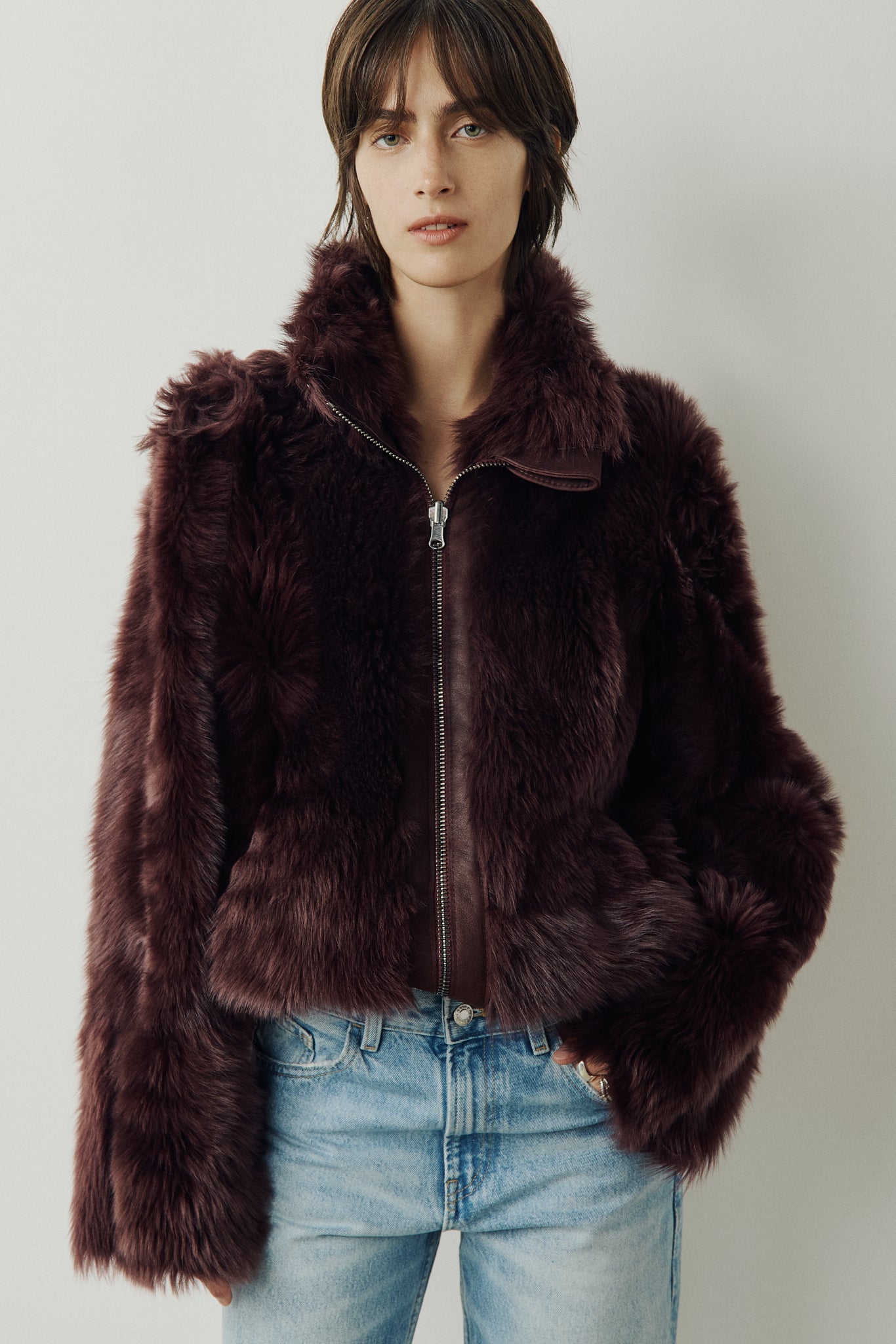 This cropped teddy jacket features a reversible design, with wool-out on one side and nappa-out on the other. The relaxed shoulder, spread collar, and slash side pockets provide comfort and functionality, while the front zip closure adds a practical touch.