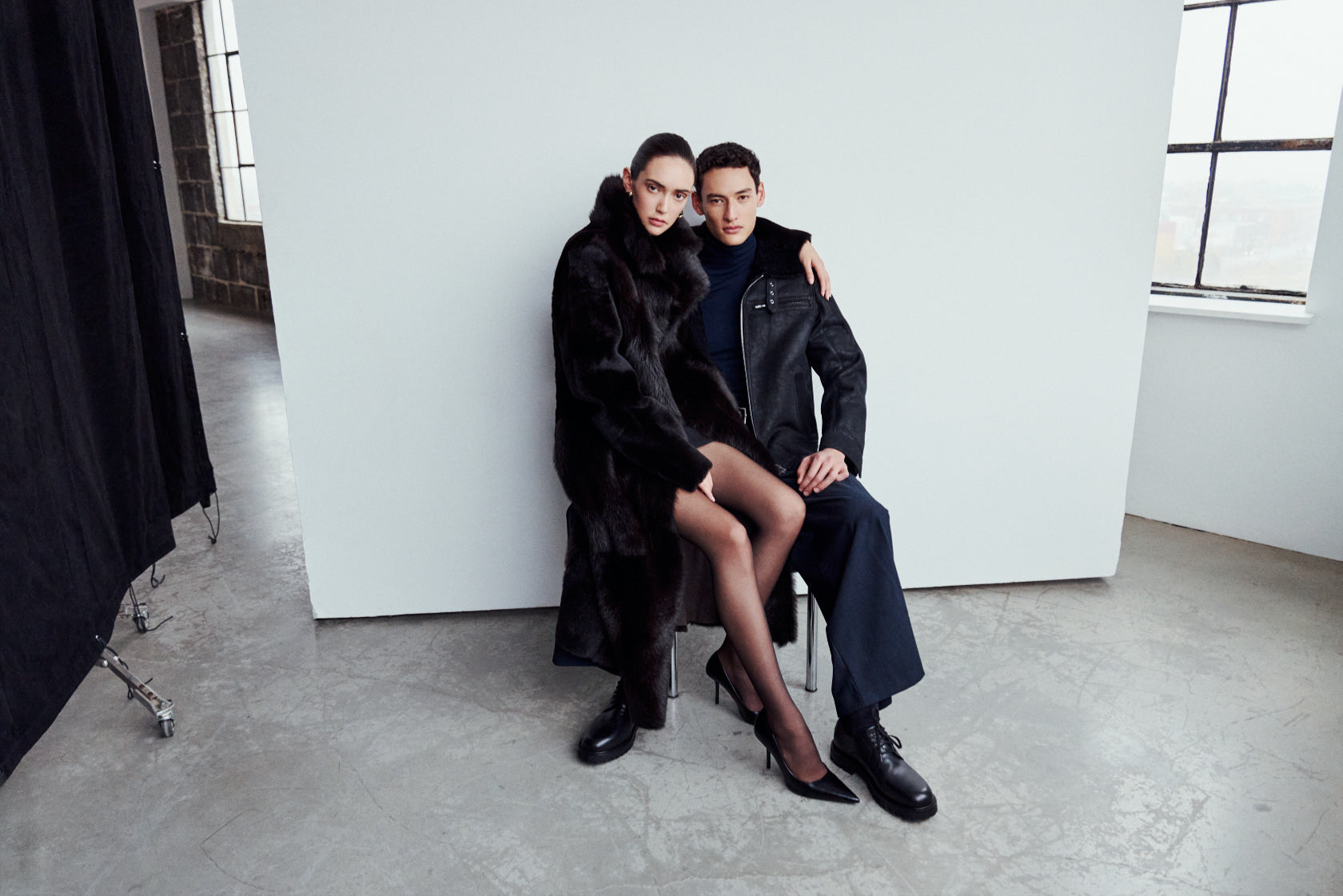 A man and woman posing together; she wears the MAEVE reversible shearling coat in chocolate suede with an oversized Toscana wool notch collar, and he wears the ARIES jet-black cracked nappa shearling moto jacket.