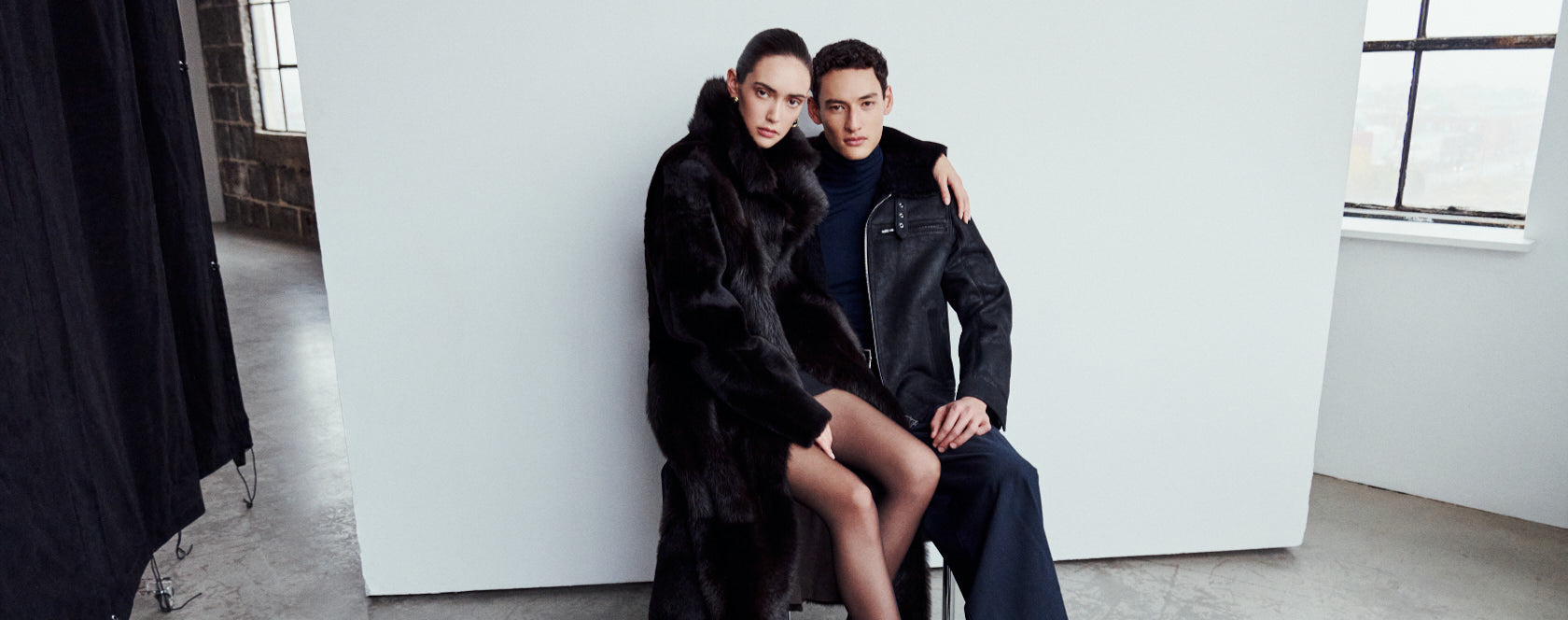 A man and woman posing together; she wears the MAEVE reversible shearling coat in chocolate suede with an oversized Toscana wool notch collar, and he wears the ARIES jet-black cracked nappa shearling moto jacket.