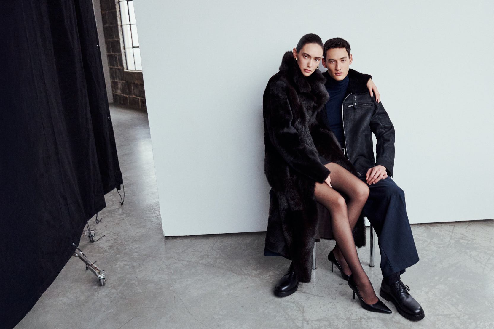 A man and woman posing together; she wears the MAEVE reversible shearling coat in chocolate suede with an oversized Toscana wool notch collar, and he wears the ARIES jet-black cracked nappa shearling moto jacket.