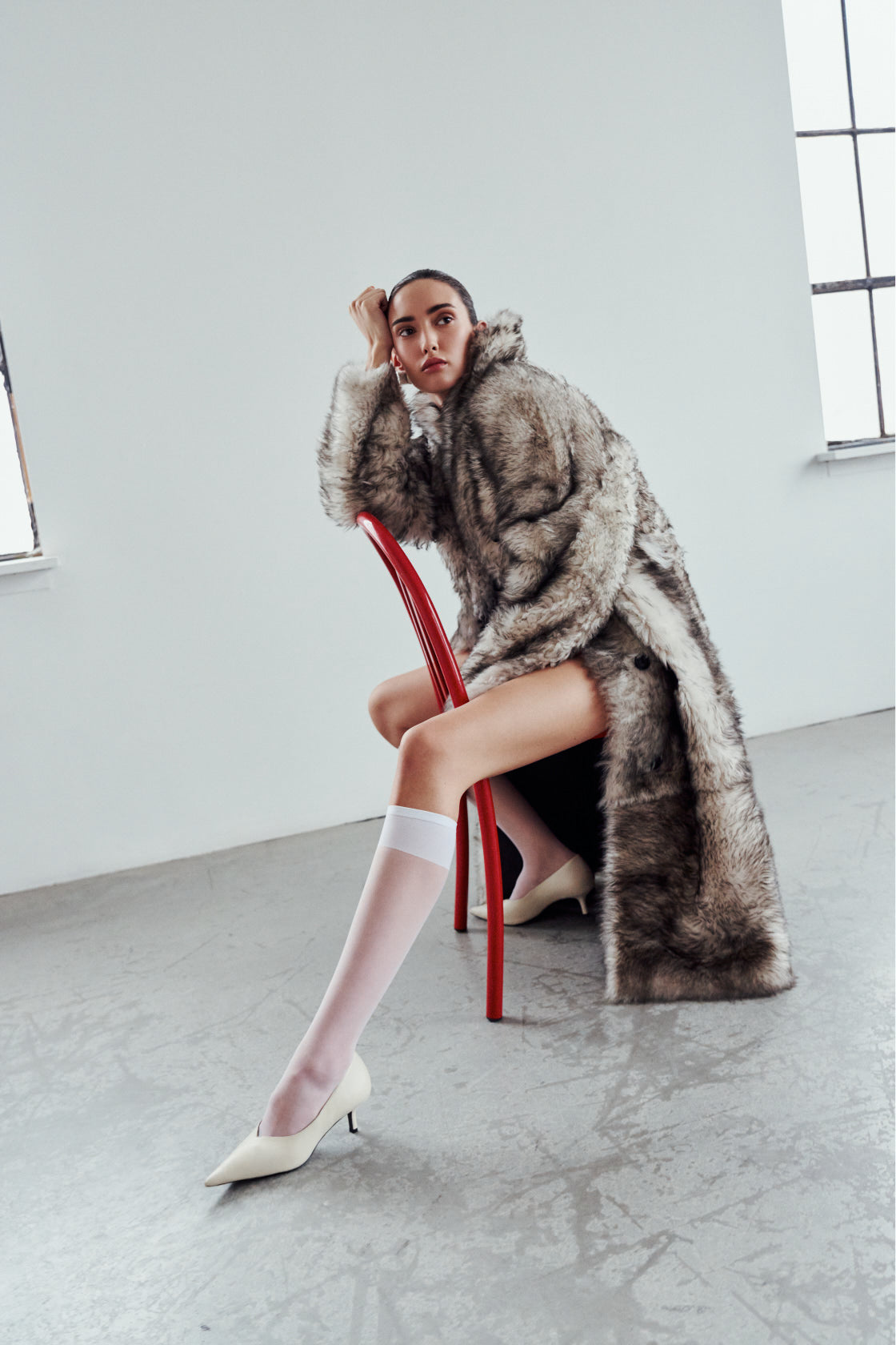 Woman wearing Magik a shearling  coat style and sitting on a red chair in a minimalistic room.