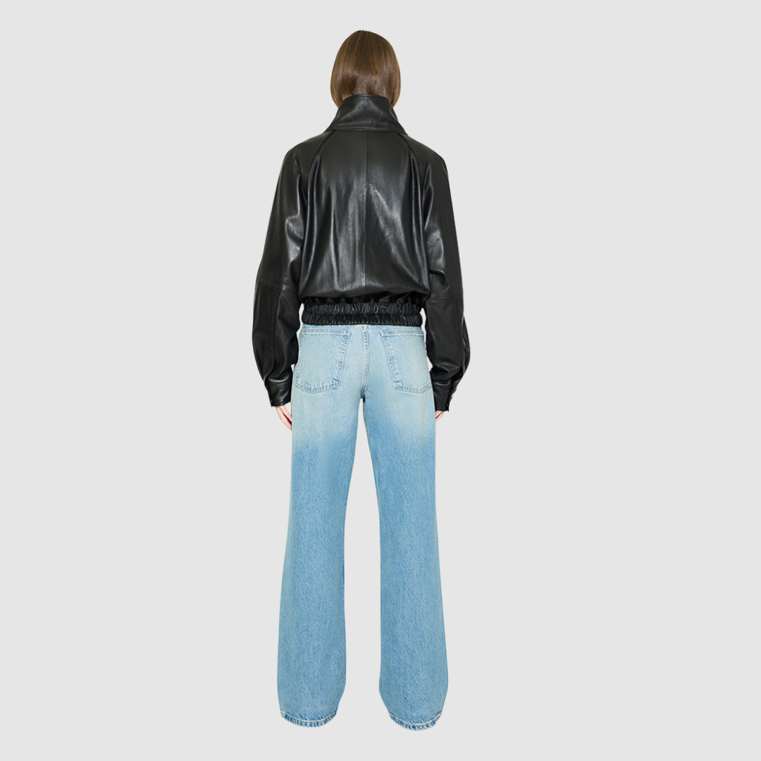 The WREN Bomber Jacket is a modern silhouette crafted in supple black nappa leather. Designed with raglan drop shoulders and an easy, relaxed volume through the body, it delivers sharp architectural presence with a sporty edge. A high stand funnel collar and concealed front zip closure with snap storm placket give it a clean, minimalist finish, while a gathered elasticized leather hem defines the waist. Fully lined for comfort, WREN serves as a definitive statement piece for transitional and cold-weather