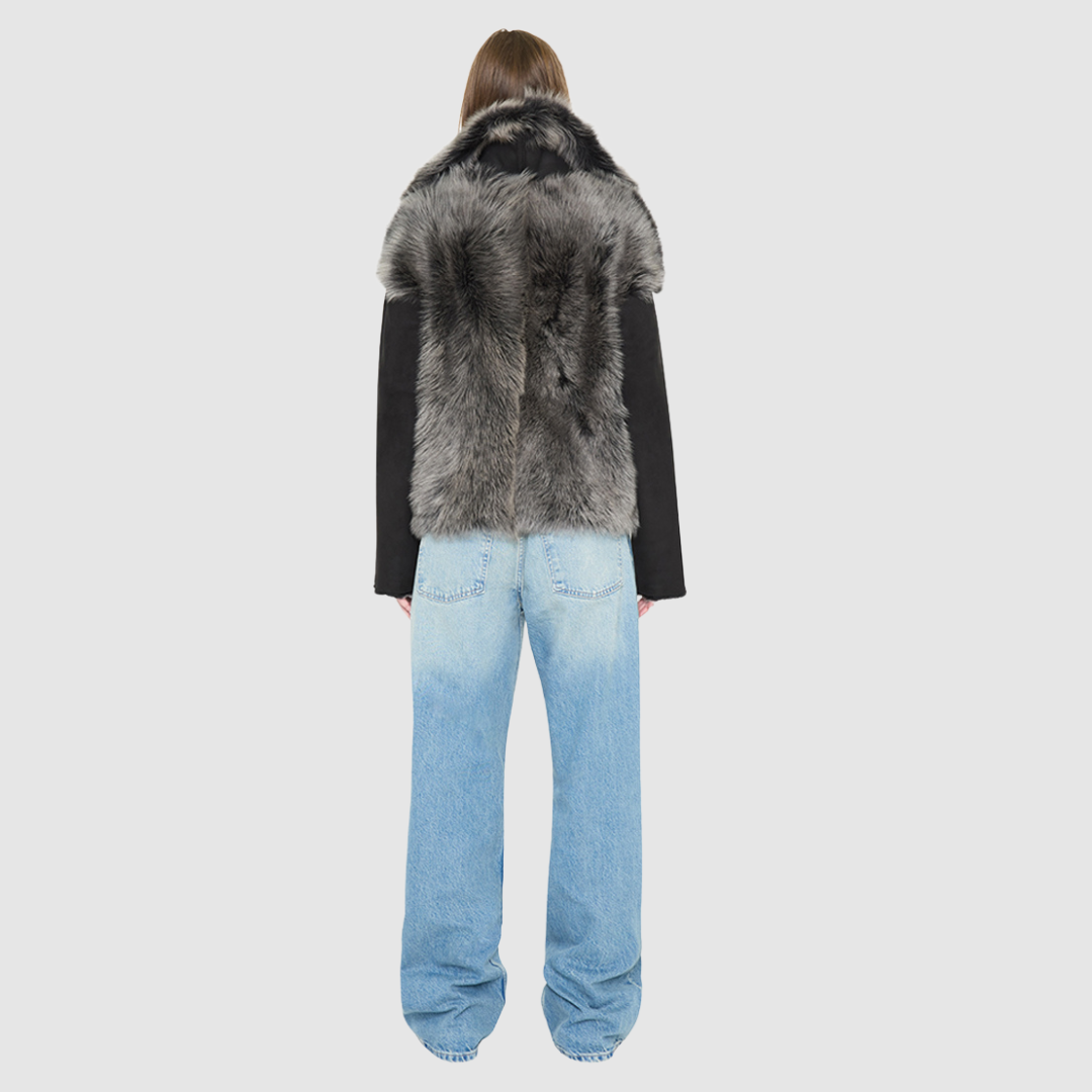 The INDIRA Shearling Jacket offers a modern take on the classic moto jacket, combining rugged function with luxurious texture. Crafted in a rich black suede that features a plush, long-haired grey Toscana shearling wool interior, it delivers an elevated cold-weather look. Designed with a distinct drop shoulder and a relaxed silhouette, it balances structure with effortless ease. Striking exposed toscana wool panels cascade down the center back and frame the front closure.