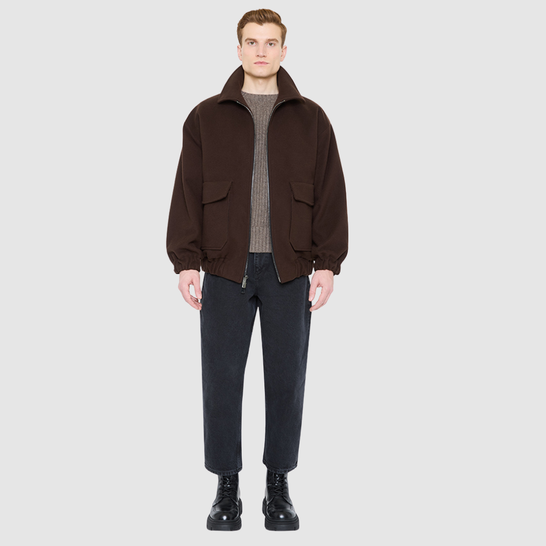 The MALCOLM Bomber Jacket is a modern silhouette crafted in a rich chocolate brown wool blend. Designed with drop shoulders and an easy, relaxed volume through the body, it delivers architectural presence and effortless style. A high convertible spread collar and two-way front zip closure give it a clean, minimalist finish, while oversized front flap patch pockets and elasticized cuffs and hem define the shape. Fully lined for comfort, MALCOLM serves as a definitive statement piece for cold-weather layering